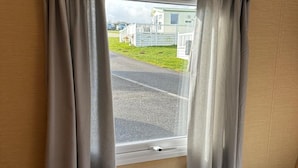 Une fenêtre avec des rideaux gris, une poignée blanche et une vue sur une route bordée d’une clôture blanche, avec une maison en arrière-plan.