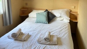Un lit bien fait, avec des draps blancs, deux serviettes enroulées et un coussin de couleur turquoise.