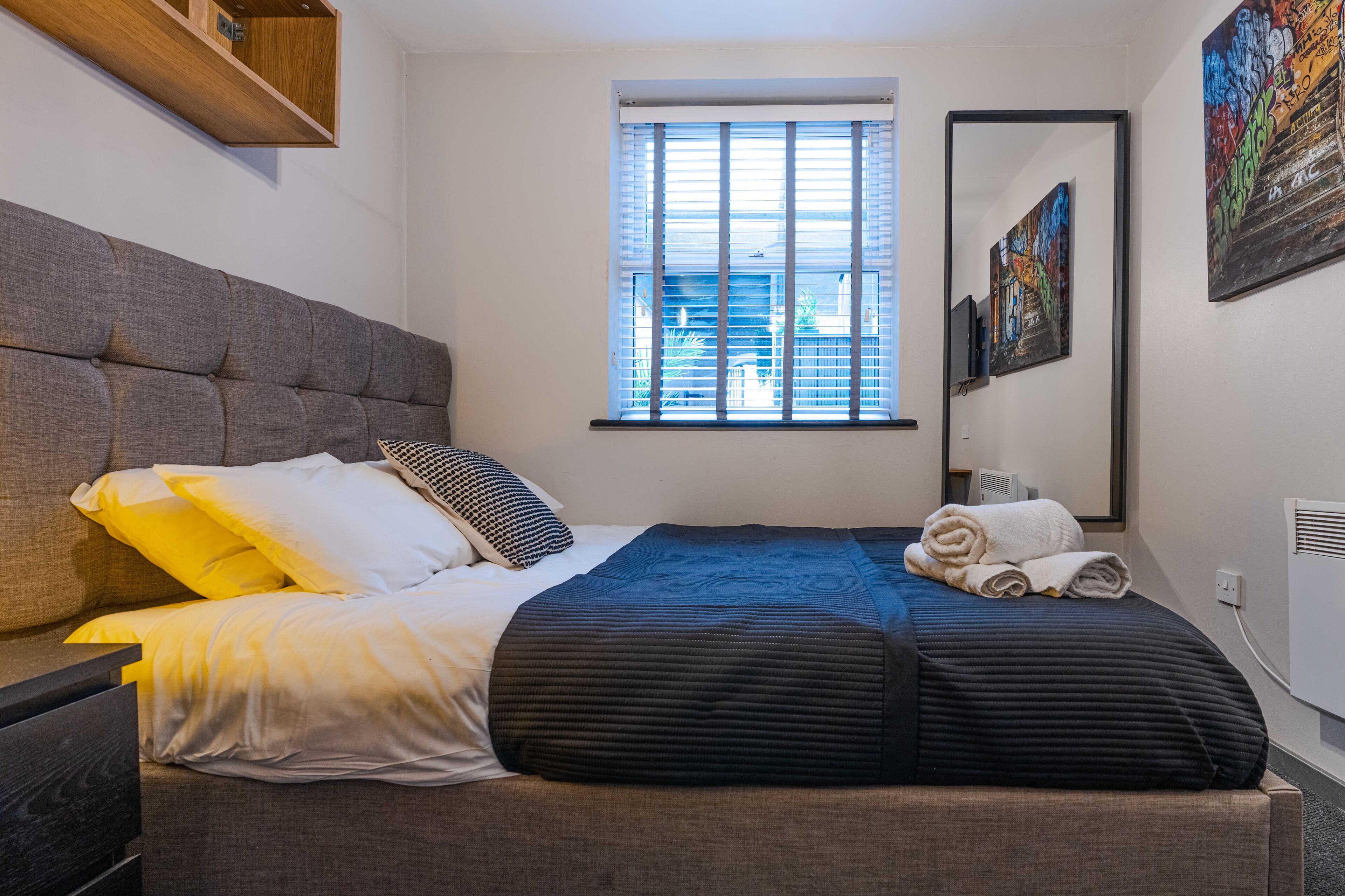 A neatly made bed with a grey headboard, a window with blinds, a mirror, and a wall with a framed artwork.
