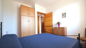 Un dormitorio con cama, armario de madera, un mueble bajo con una planta y una puerta que da a otra habitación.