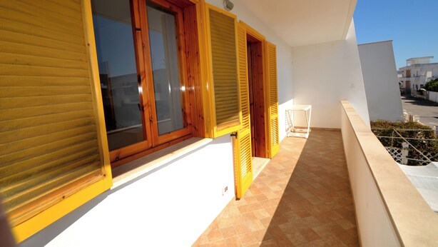Un balcón con contraventanas de madera, una silla blanca y vista a la calle.