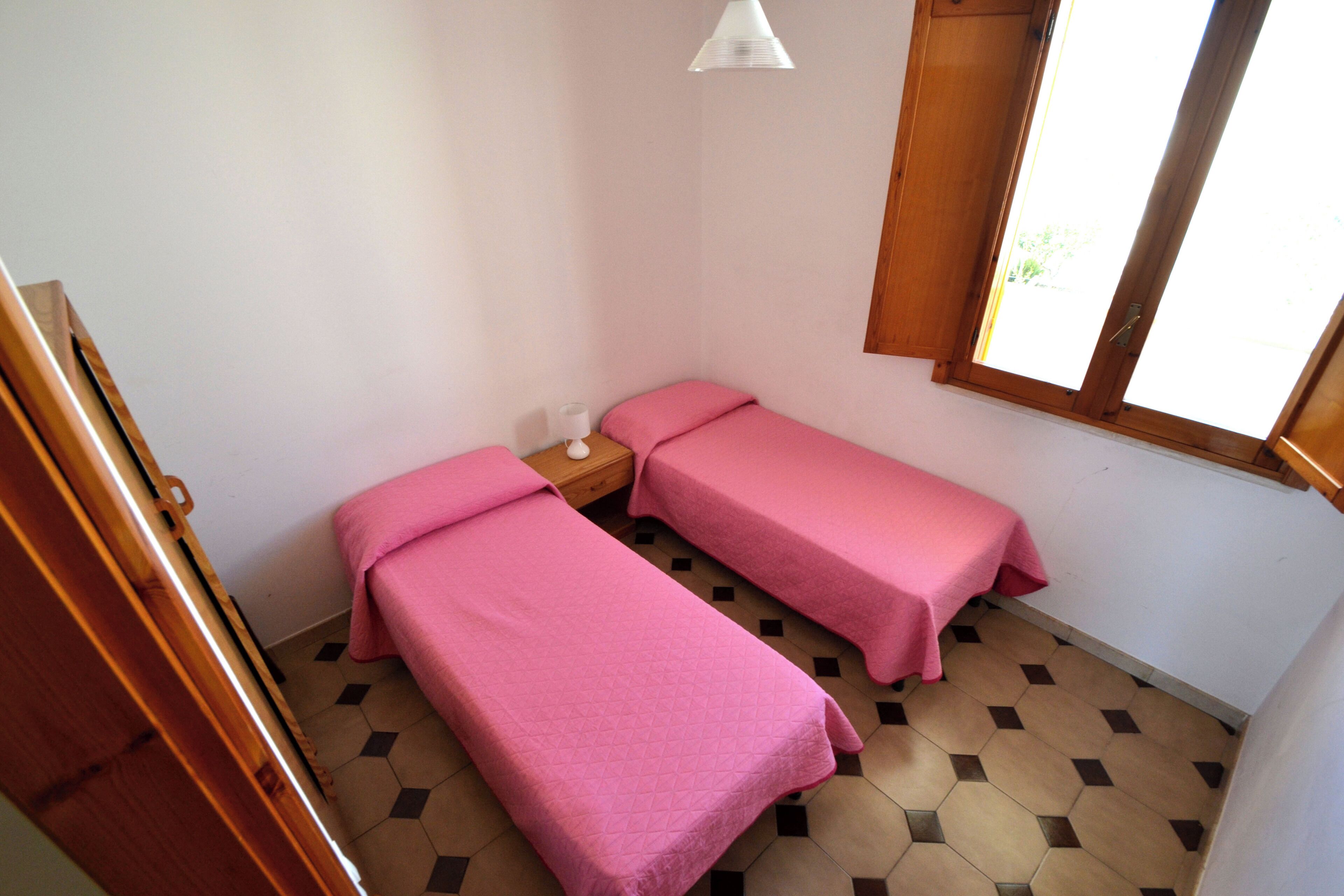 A small room with two beds, a wooden wardrobe, a bedside table with a lamp, and a window with a view.
