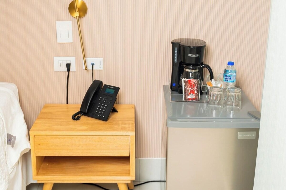 A compact hotel room with a wooden nightstand, a phone, a coffee machine, and a water dispenser.