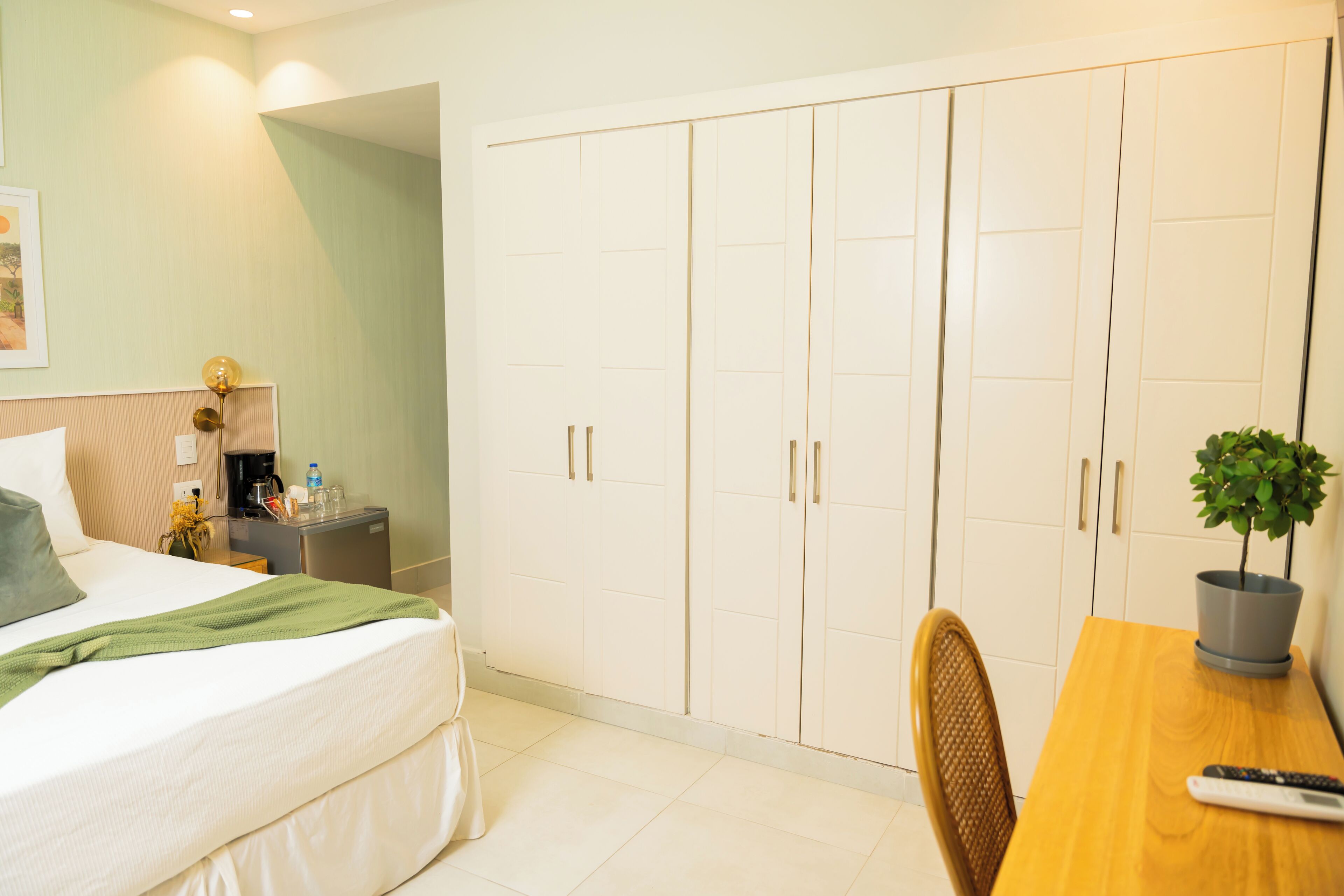 Una habitación de hotel con una cama, un escritorio con una silla, una planta en maceta y un armario.