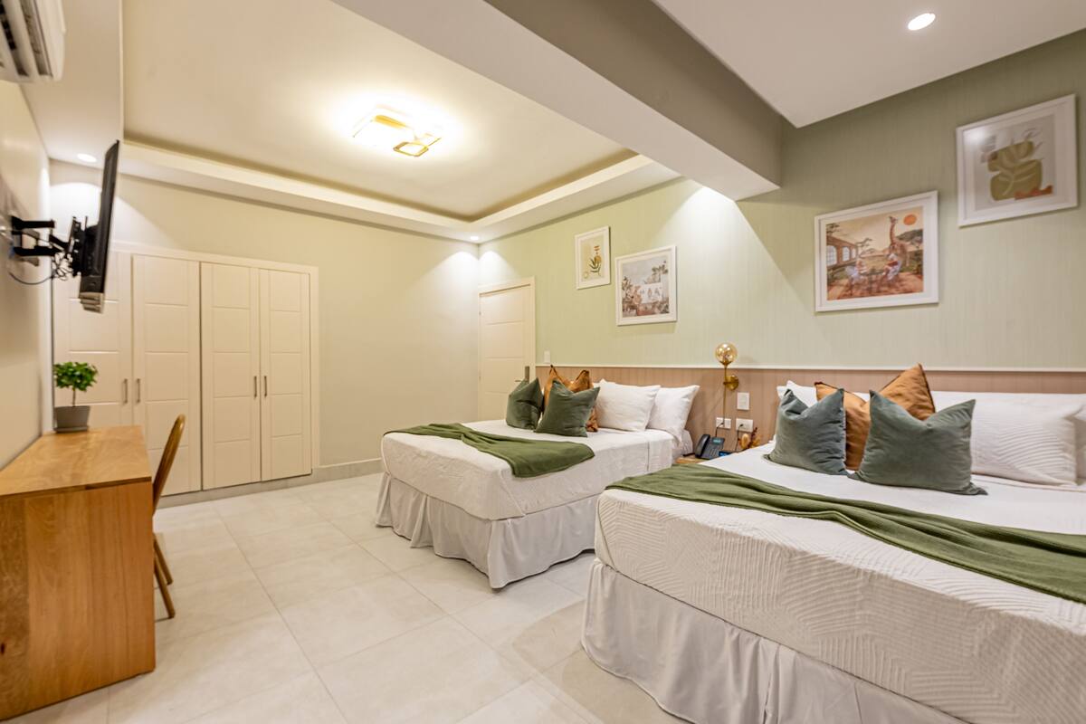A hotel room with two beds, a desk, and a chair. There are wall-mounted air conditioning units and a ceiling light fixture.