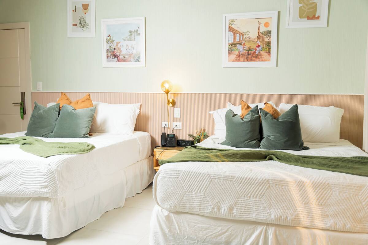 A hotel room with two beds, each with a white bedspread and pillows, a bedside table with a lamp, and framed pictures on the wall.