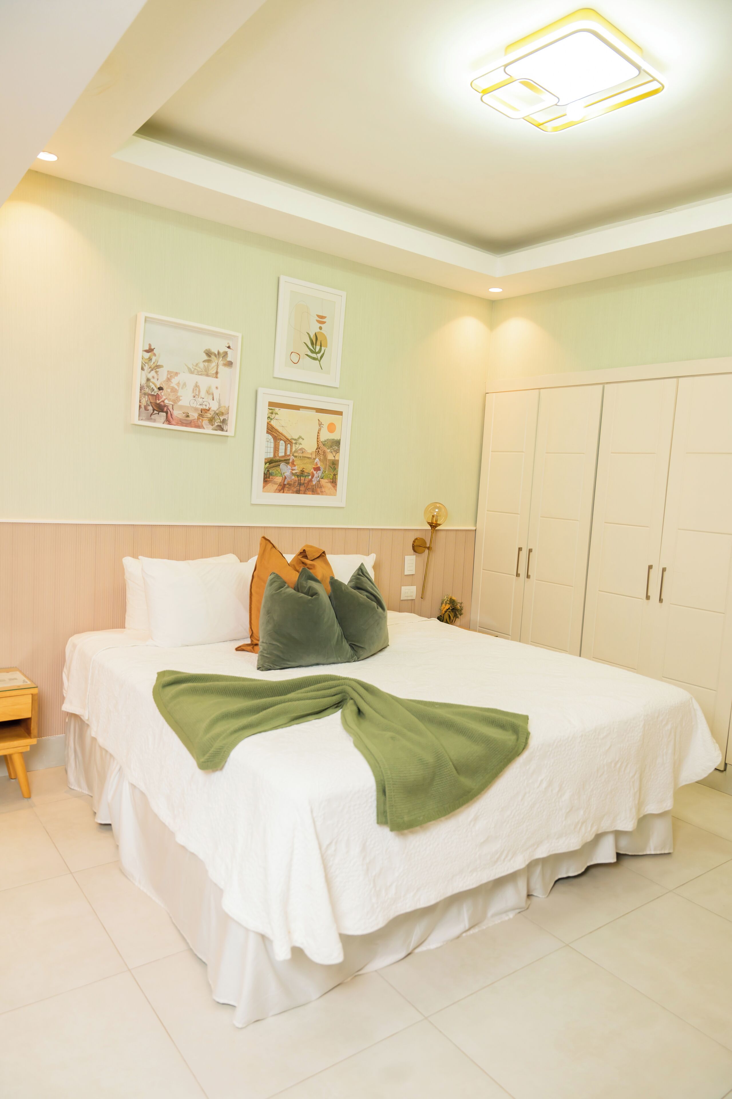 Uma cama bem arrumada com colcha branca e verde, uma mesa de cabeceira de madeira e um guarda-roupa com duas portas.