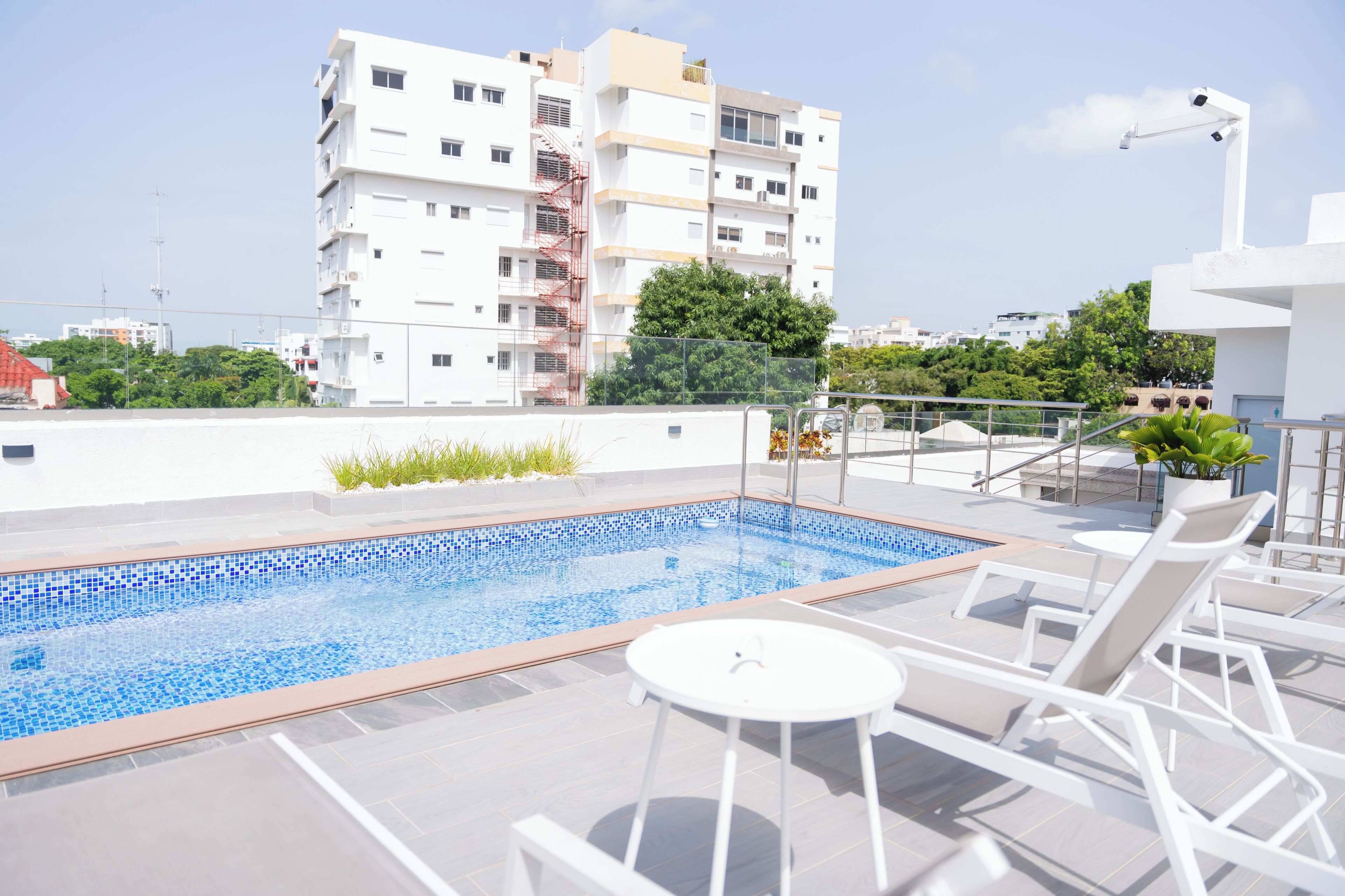 A rooftop swimming pool with a tiled edge, surrounded by white lounge chairs and a small table.
