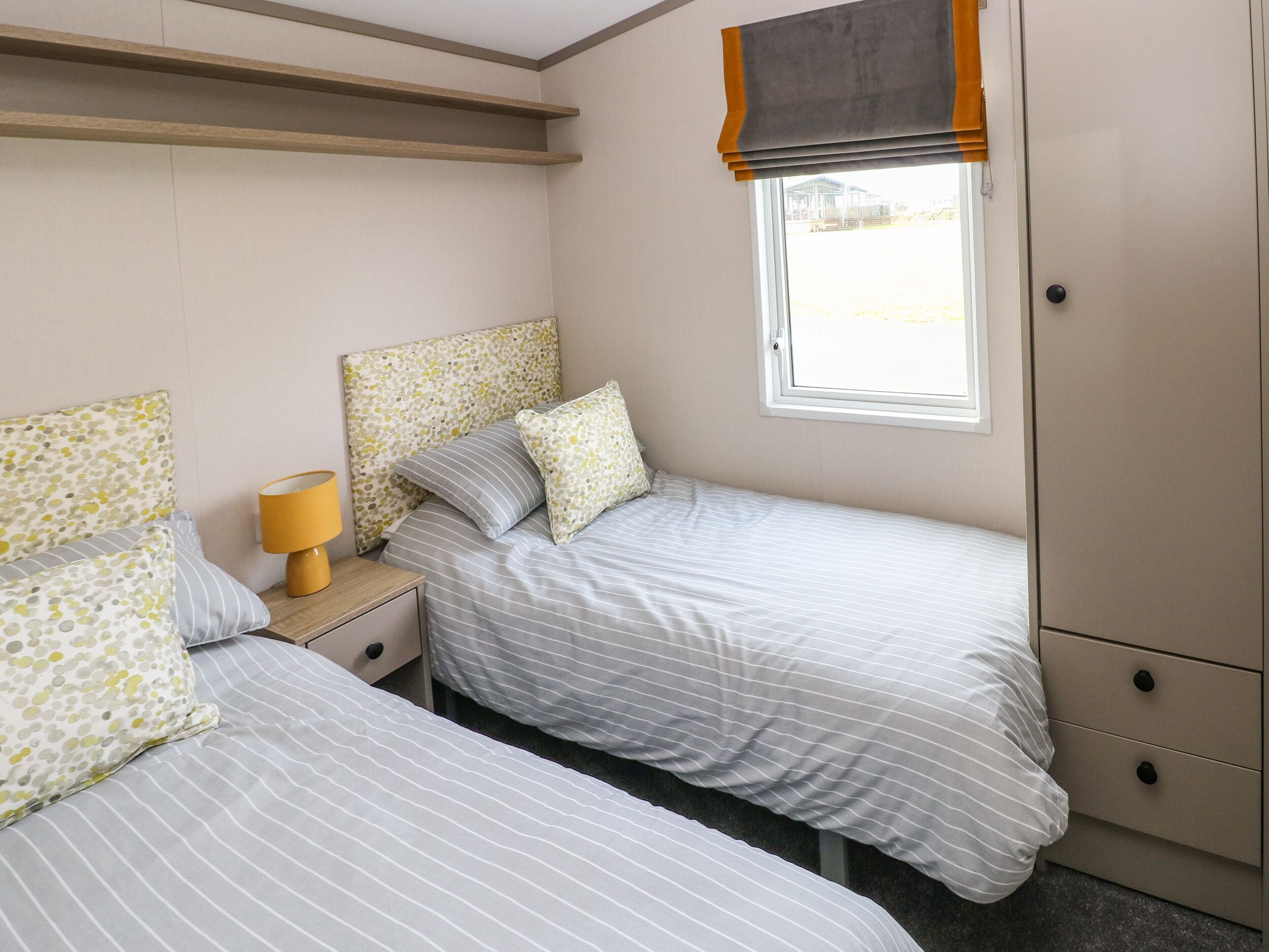 A bedroom with two beds, a nightstand with a lamp, a window with a blind, and a wardrobe.