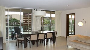 A modern living room with a dining table, chairs, a sofa, and a large window with a view of palm trees. - Herdade dos Salgados, T3 8D-0B, Vila das Lagoas (Albufeira)