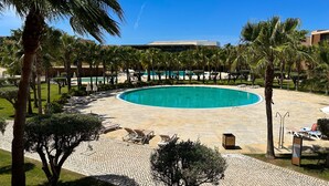 A circular pool with lounge chairs and palm trees, surrounded by a paved area and a building in the background. - Herdade dos Salgados, T3 8D-0B, Vila das Lagoas (Albufeira)