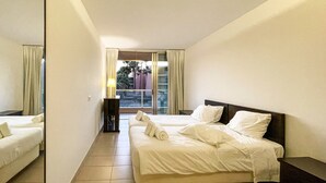A hotel room with two beds, a large window with curtains, a bedside table, and a view of palm trees outside. - Herdade dos Salgados, T3 8C-1B, Vila das Lagoas (Albufeira)