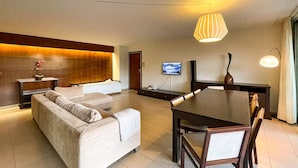 A modern hotel room with a sofa, a dining table with chairs, a TV mounted on the wall, and a large window. - Herdade dos Salgados, T3 8C-1B, Vila das Lagoas (Albufeira)