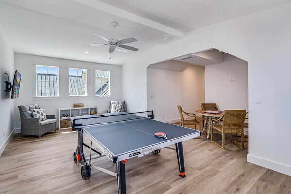 A spacious room with a ping pong table, a ceiling fan, and a dining area with wicker chairs.