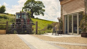 A rural property with a large tree, a barn, and a paved driveway leading to a building with large windows. - Luxury 2-Bed Cotswolds Barn with Private Hot Tub | Sleeps 4 (Gloucester)
