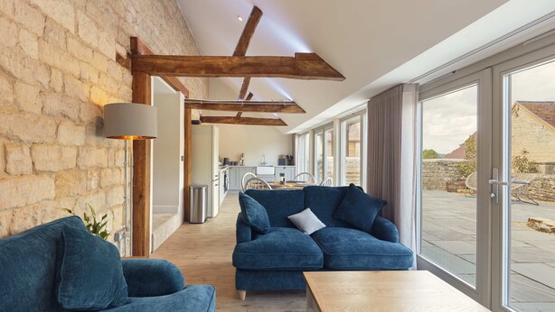 A modern living room with a blue sofa, a wooden coffee table, and a large window offering a view of the outside. - Luxury 2-Bed Cotswolds Barn with Private Hot Tub | Sleeps 4 (Gloucester)