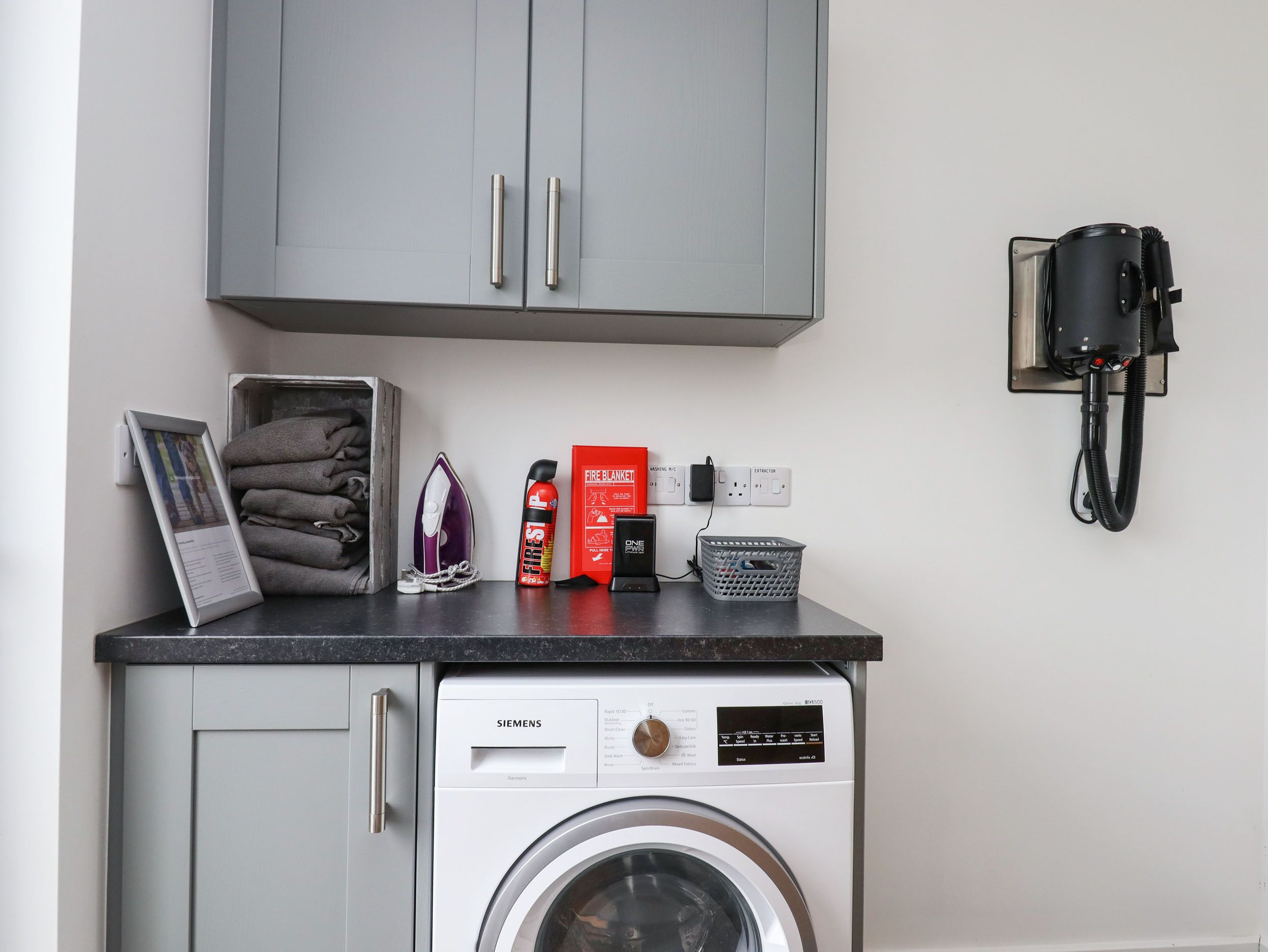 A compact laundry area with a Siemens washing machine, a stack of folded towels, a fire blanket, an iron, and a vacuum cleaner.