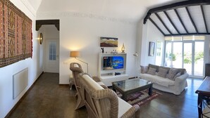 A spacious living area with a white sofa, wicker chairs, a glass coffee table, and a wooden dining table. A television is mounted on the wall, and there is a large window providing natural light. - Casa Saskia with view to Avocado plantation and Atlantic.
Heated Poolarea (La Matanza de Acentejo)