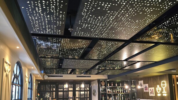 A restaurant interior with a modern ceiling design, wooden furniture, and a bar area. - Hotel Apricia  (Ludhiana)