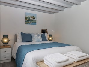 A neatly made bed with blue bedding, two bedside lamps, a painting of a coastal scene, and a stack of white towels on a wooden bench.