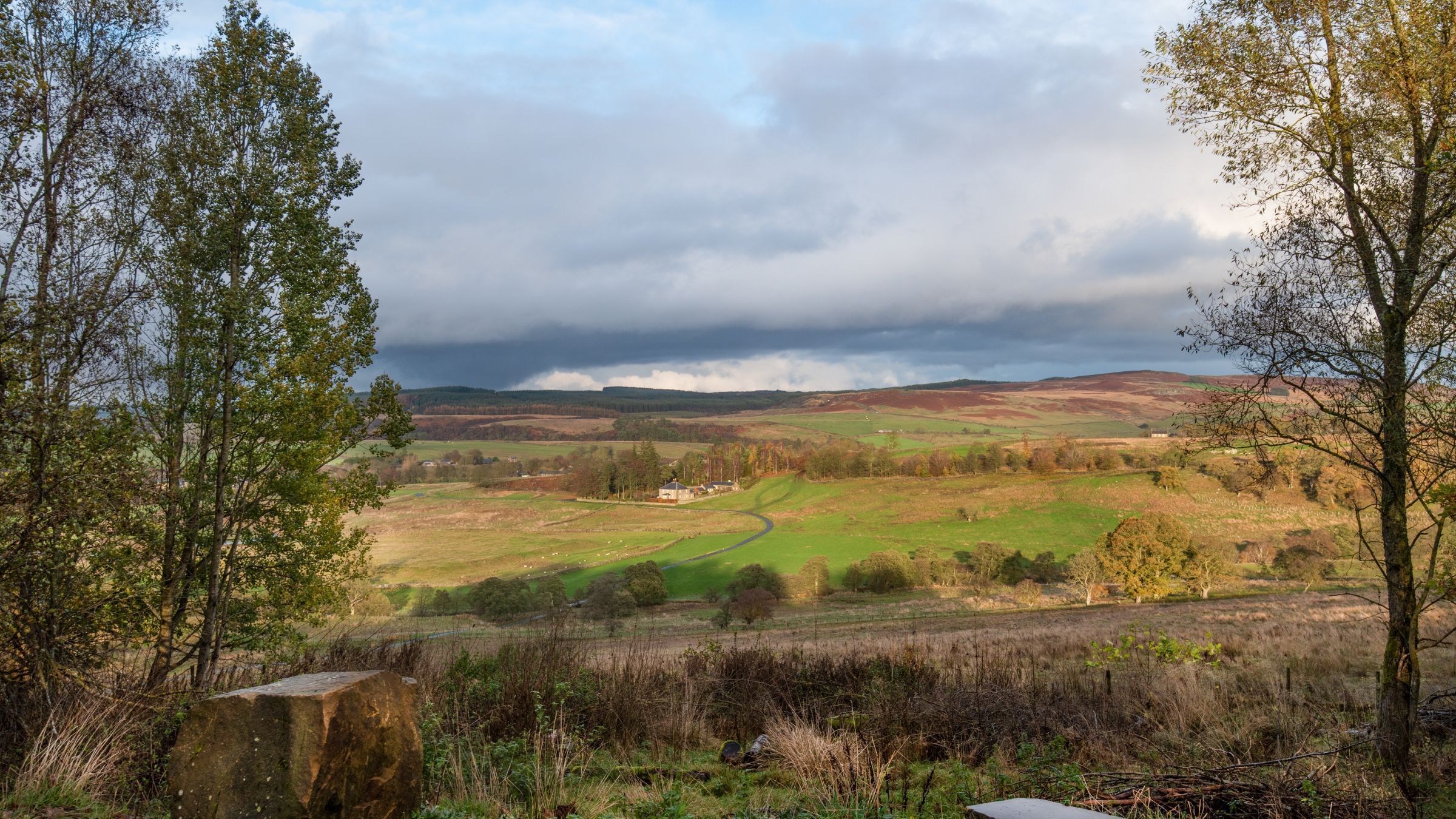 Un paisaje rural con colinas onduladas, una casa pequeña y un muro de piedra.