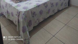 A single bed with a floral bedspread, a white pillow, and a rolled towel on the bed. There is a small table with a cloth and an iron on it. The room has plain walls and tiled flooring. - Apartment, Bairro Santa Lúcia, covered garage 2.50x4.70 (Montes Claros)