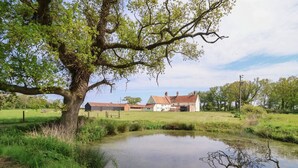 Un paysage rural avec un étang, un grand arbre et une ferme dont le toit est recouvert de tuiles rouges.