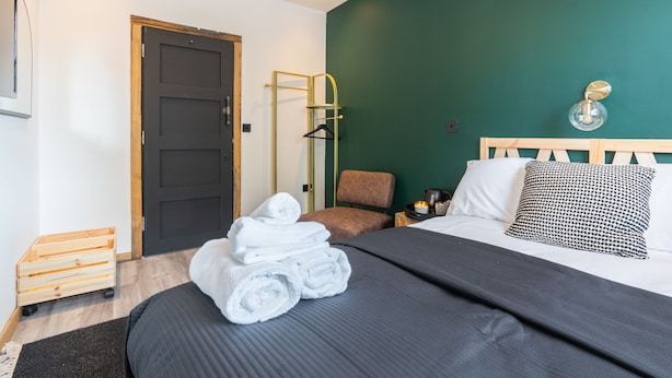 Une chambre d’hôtel avec un lit, une chaise, une table d’appoint en bois, un luminaire suspendu et une porte.