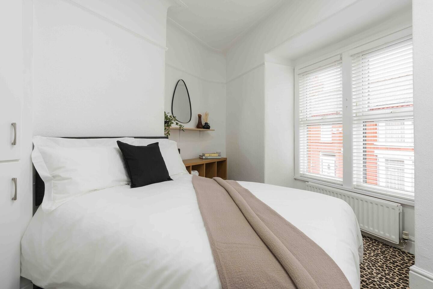 A neatly made bed with white bedding and a brown throw, a small bedside table with a book, and a window with blinds.