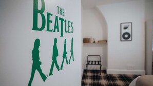 Une chambre avec un motif décoratif représentant les Beatles sur un mur, un lit recouvert d’une couverture à carreaux, une table de chevet et un tableau encadré accroché au mur.