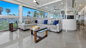 Ein modernes Wohnzimmer mit einem Glastisch, einem weißen Sofa mit gemusterten Kissen und großen Fenstern mit Blick auf Palmen.