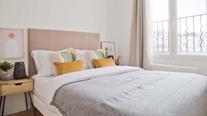 A neatly made bed with a grey blanket, a bedside table with a lamp, and a window with a view of buildings. - Trendy familly apt in Mouffetard (Paris)