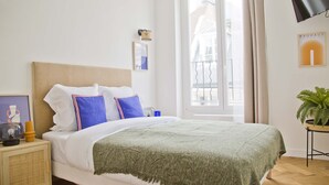 A neatly made bed with white bedding and a green throw, a bedside table with a lamp and a picture frame, a potted plant, and a window with curtains. - Trendy familly apt in Mouffetard (Paris)