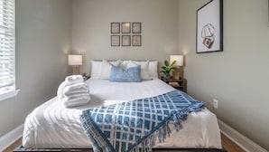 Una cama bien hecha con un plaid azul, una pila de toallas blancas y una mesita de noche con una lámpara.