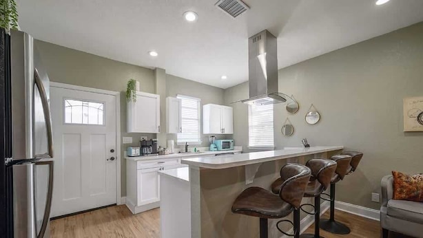 Una cocina moderna con armarios blancos, una isla central con taburetes de bar, un refrigerador de acero inoxidable y un microondas.