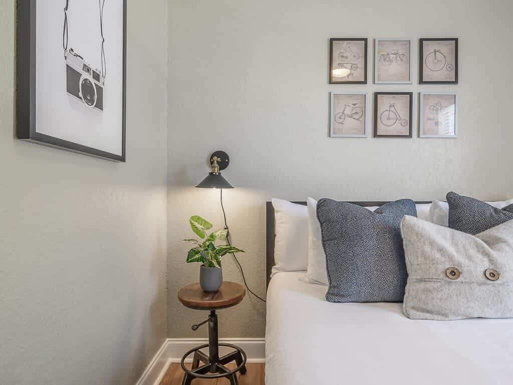 A bedroom with a bed, pillows, a small table with a plant, a lamp, and framed pictures on the wall.