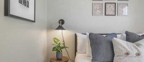 A bedroom with a bed, pillows, a small table with a plant, a lamp, and framed pictures on the wall.