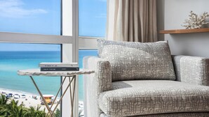 Ein Zimmer mit Blick auf den Strand, ein Sessel, ein kleiner Tisch mit Büchern und ein Regal mit einem Dekorationsobjekt.