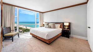 Ein Hotelzimmer mit einem großen Bett, einem Sessel, einem Nachttisch mit Lampe und Blick auf Strand und Meer.