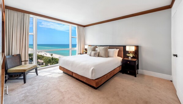 Habitación de hotel con una cama grande, una silla, una mesita de noche con lámpara y vistas a la playa y al océano.