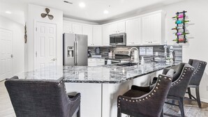Una cocina moderna con armarios blancos, encimera de granito y horno empotrado. Hay dos taburetes de bar con hebillas decorativas.