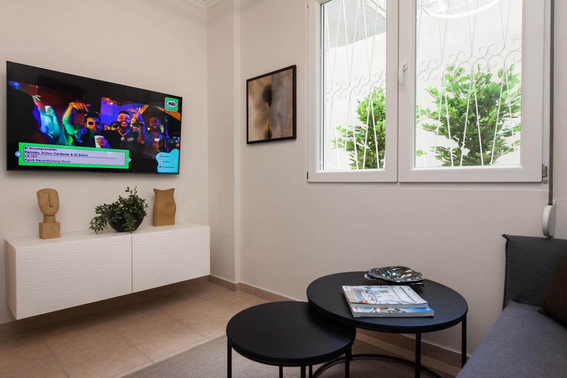 Un saló modern amb televisió de pantalla plana penjada a la paret, una taula petita amb revistes, un sofà i una finestra amb vista a vegetació.