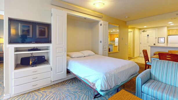 A hotel room with a bed, a TV on a stand, a desk with a chair, and a kitchenette.