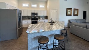 A modern kitchen with white cabinets, a stainless steel refrigerator, a microwave, and a built-in oven. There is a marble island with two stools and a sink. - Fabulous 4 bedroom, 3 bath home in gate community of Briarwood (Naples)