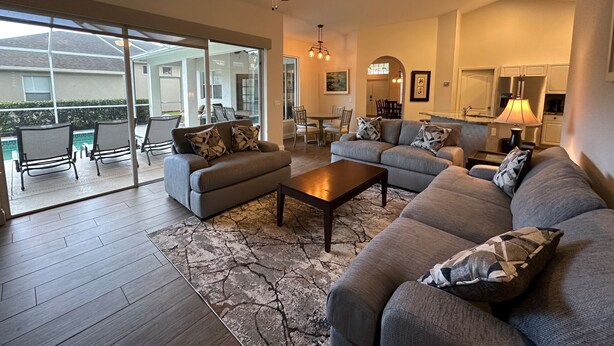 A spacious living room with a large gray sectional sofa, a coffee table, and a ceiling fan. There is a sliding glass door leading to an outdoor patio area with chairs and a dining table. - Fabulous 4 bedroom, 3 bath home in gate community of Briarwood (Naples)