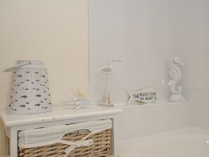 A bathroom with a white tiled wall, a white shelf with decorative items, a white towel rack, and a wicker basket. - Woodmans Cottage (Falmouth)