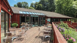 Eine Außenpatio mit Holztischen und Metallstühlen, ein rotes Gebäude mit einem Schild, auf dem "Pendra Loweth Bar" steht, und ein Holzdeck mit Geländern und Topfpflanzen.