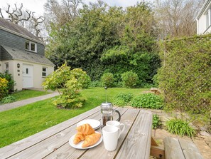 A wooden picnic table with croissants, a coffee pot, and mugs in a garden with a house in the background. - Woodmans Cottage (Falmouth)