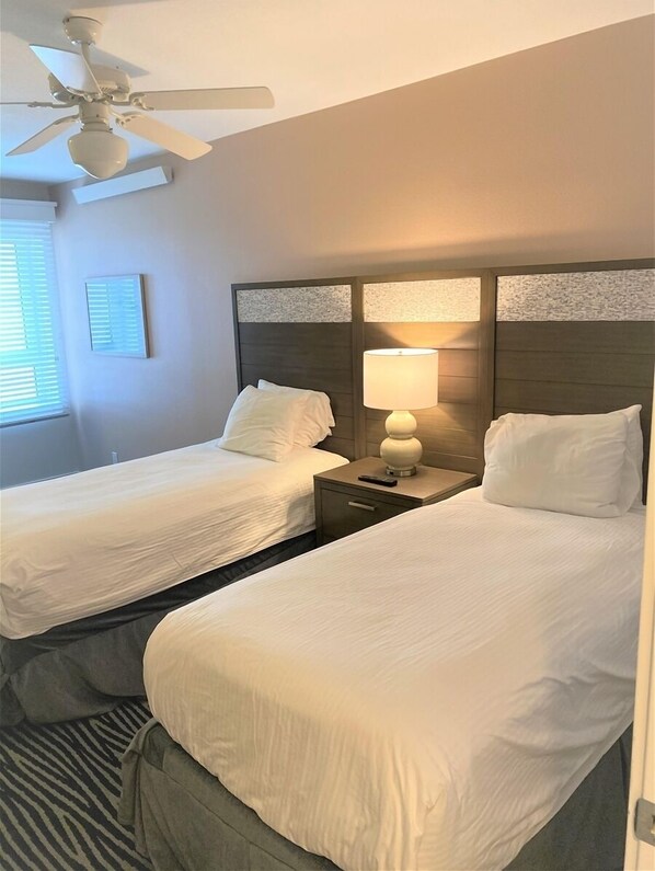 A hotel room with two beds, a ceiling fan, a nightstand with a lamp, and a window with blinds. - Seaside Resort - Ocean Front 3 Bedroom 2 Bath - Assignment 1 (Seaside)