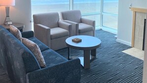 Quarto de hotel moderno com uma janela grande que oferece vista para o oceano, um ventilador de teto, um sofá, uma poltrona e uma pequena mesa.